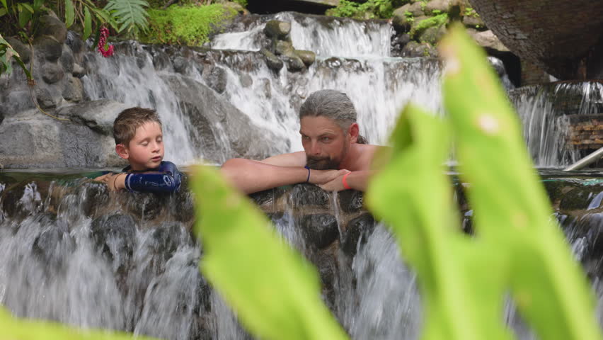 Father and son relax together in a tropical hot springs pool surrounded by lush green foliage. Water cascades over rocks as they lean on the stone edge enjoying quality time.
