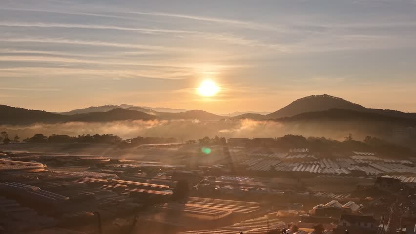 Drone zoom shot over massive industrial greenhouses during a vibrant sunrise. The sunburst is visible on the horizon, bathing the entire scene in golden light and casting long shadows in the valley.
