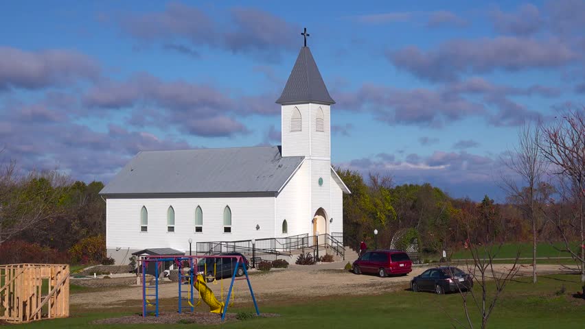 Scenic view of a white country church with a grey steeple, featuring a playground and parked cars under a blue sky with clouds. Autumn trees in background.