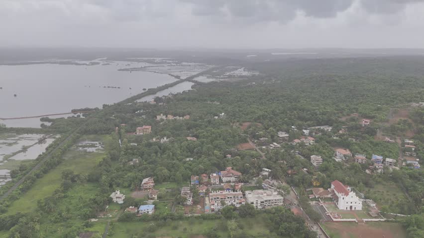 Aerial view of Old Goa and the Mandovi River during the monsoon season, featuring coconut palms, green fields, backwaters, lush greenery, nearby houses, and cloudy skies.