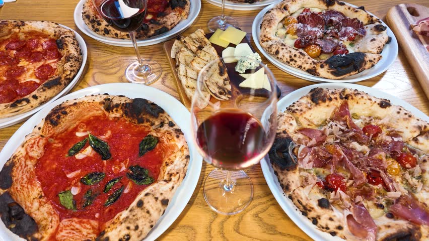 High-angle shot of multiple pizzas, cheese board, and red wine on a wooden table.
