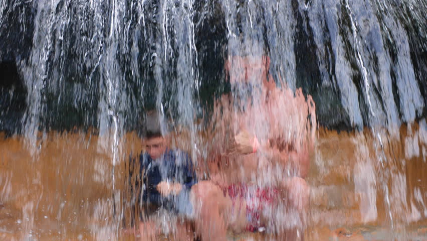 Father with son under waterfall in slow motion as cascading water splashes over happy pair. Man and boy give thumbs up gestures while enjoying refreshing summer adventure together outdoors.