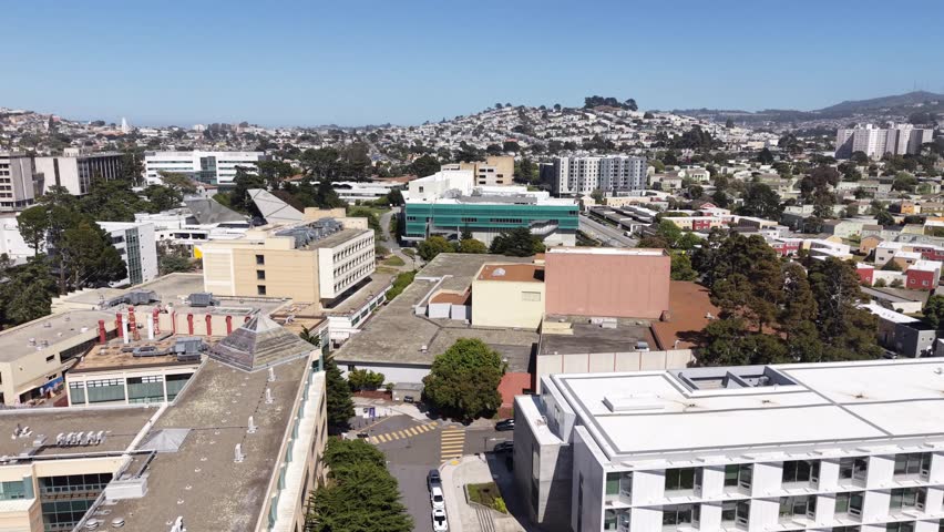 Backward drone reveal of the San Francisco State University campus buildings and surrounding neighborhood on a bright, sunny day with clear blue skies showing the urban landscape and distant hills