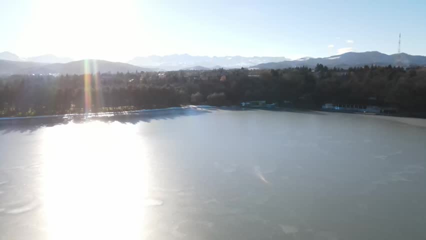 Aerial drone pull-back over a frozen pond covered with snow and ice, with bright winter sunlight reflecting on the surface and forested mountain slopes in the background