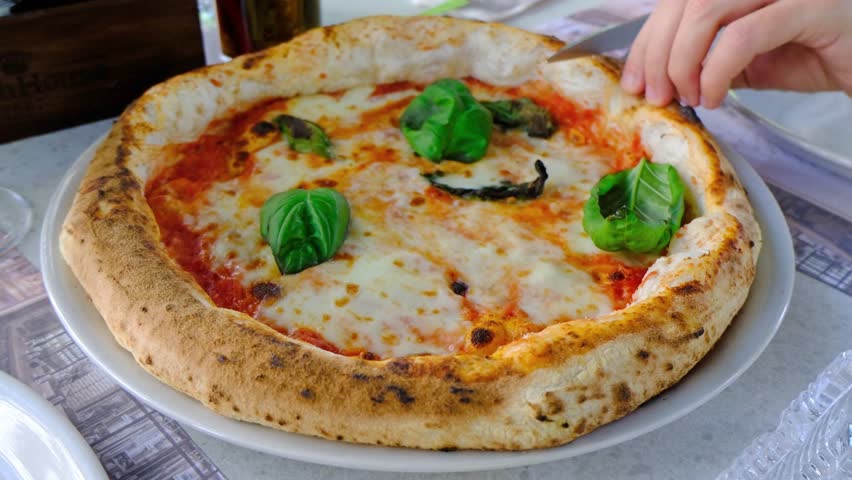 A man cuts a pizza with scissors. The pizza has a golden crust, topped with cheese and fresh basil leaves. The setting is casual and inviting.