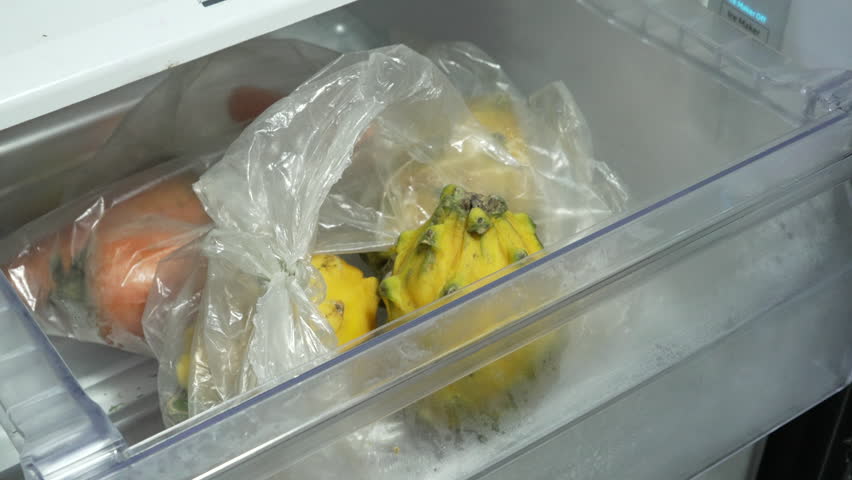A person reaches into a fridge crisper drawer and lifts a yellow dragon fruit from a plastic bag among other stored produce.