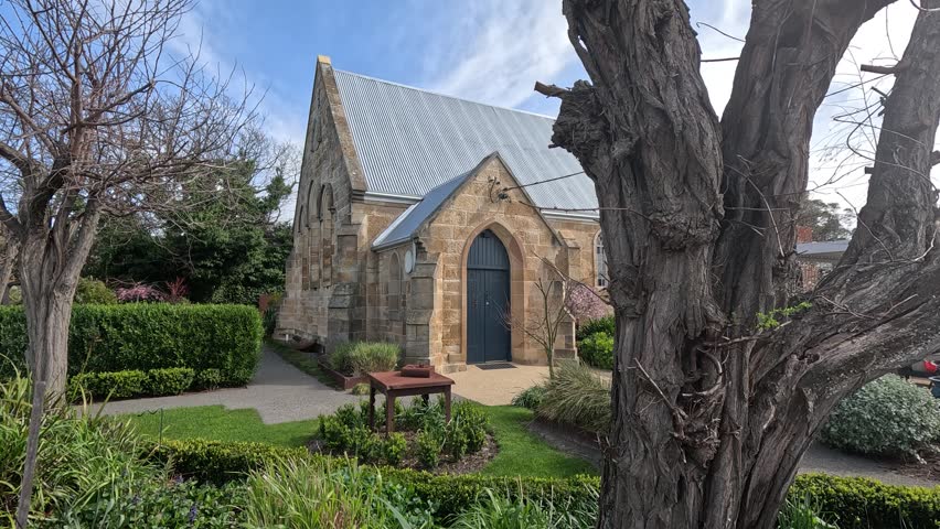A leafless tree frames a stone chapel with arched blue door and manicured garden paths on a clear winter day.