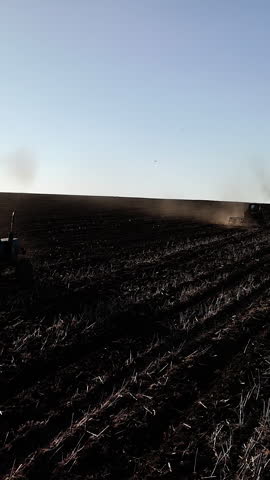 Tractor plowing a field in a rural landscape, creating dust clouds as it moves through rows of freshly tilled soil under a clear blue sky