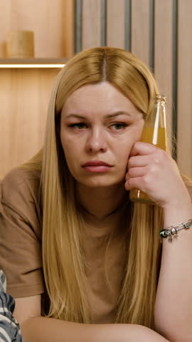 Young Caucasian woman with long blonde hair rests her head on her hand while holding a drink, seated at a table with wooden background and soft lighting