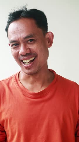 Asian man with an ugly funny face wearing an orange t-shirt smiles cheerfully against a plain white background