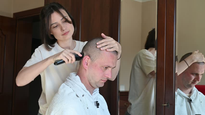 A woman gives her husband a very short buzz cut using electric hair clippers in a room at home. The video captures a casual home haircut, family life, personal grooming, and everyday domestic activities in a relaxed indoor setting.