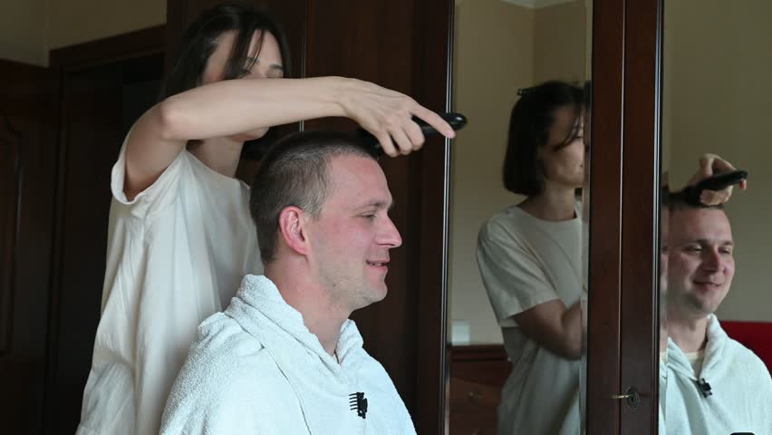 A woman gives her husband a very short buzz cut using electric hair clippers in a room at home. The video captures a casual home haircut, family life, personal grooming, and everyday domestic activities in a relaxed indoor setting.