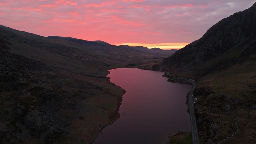 A drone scenery of Llyn Ogwen lake with A5 road along the shoreline between mountains in Snowdonia National Park, Wales during sunrise with pink sky