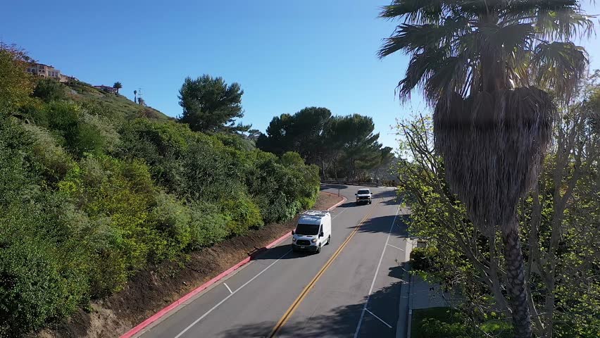 Aerial view of cars driving on a scenic California hillside road past lush green trees and a palm tree under a clear blue sky.