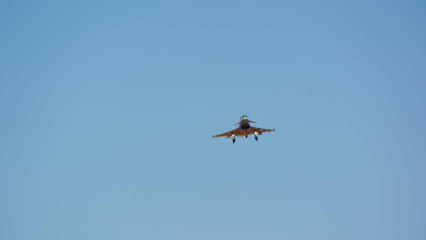 Military fighter jet on approach descending low over an airfield perimeter fence and dry field, combat aircraft coming in to land above golden grass, warplane inbound