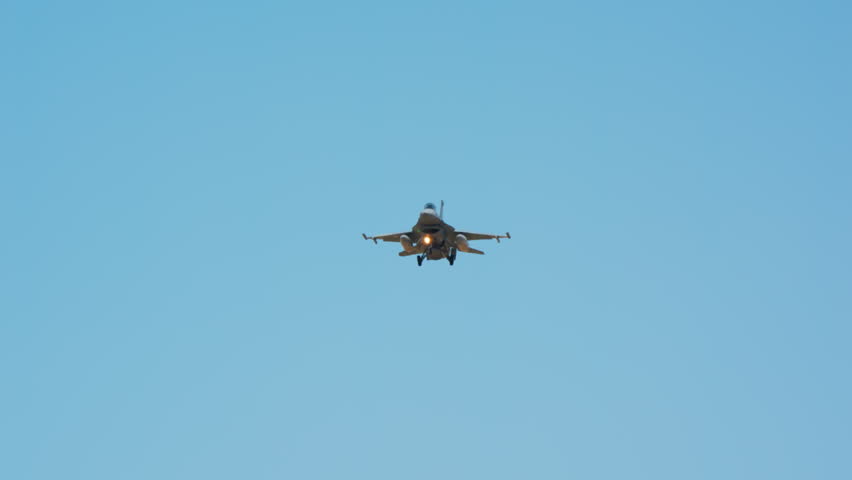 Military fighter jet on approach seen head on with gear down and landing light on, combat aircraft descending against blue sky, warplane coming in low to land