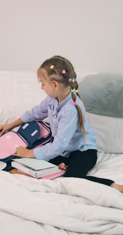 Little schoolgirl packing her notebooks and books into a backpack on bed.