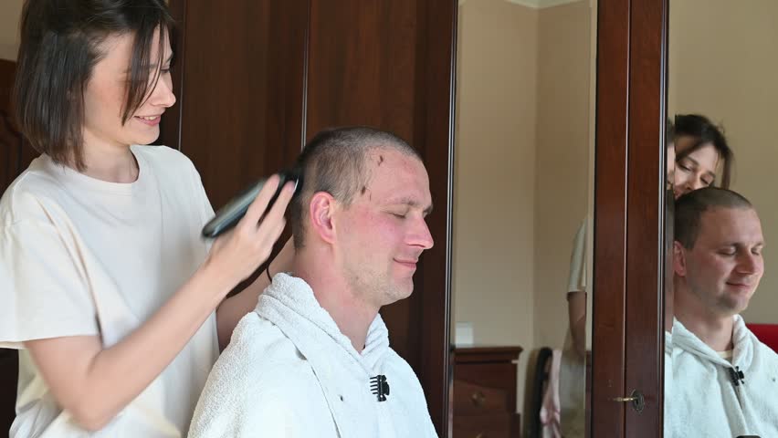 A woman gives her husband a very short buzz cut using electric hair clippers in a room at home. The video captures a casual home haircut, family life, personal grooming, and everyday domestic activities in a relaxed indoor setting.
