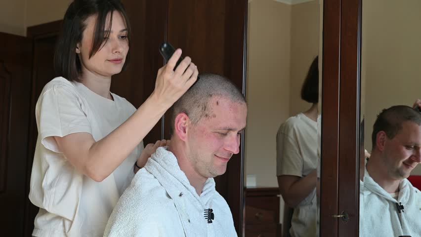 A woman gives her husband a very short buzz cut using electric hair clippers in a room at home. The video captures a casual home haircut, family life, personal grooming, and everyday domestic activities in a relaxed indoor setting.