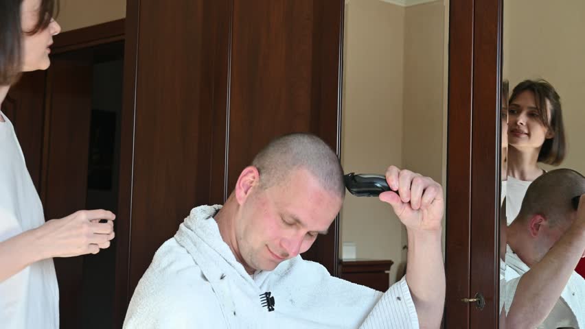 A woman gives her husband a very short buzz cut using electric hair clippers in a room at home. The video captures a casual home haircut, family life, personal grooming, and everyday domestic activities in a relaxed indoor setting.