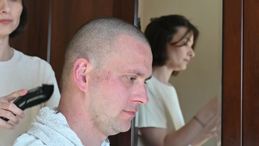 A woman gives her husband a very short buzz cut using electric hair clippers in a room at home. The video captures a casual home haircut, family life, personal grooming, and everyday domestic activities in a relaxed indoor setting.