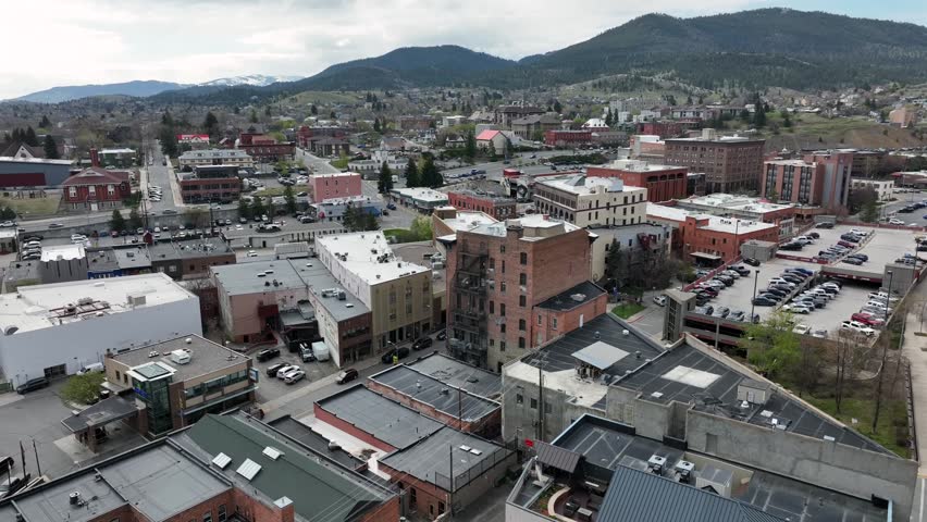 Helena’s downtown mixes historic brick buildings and modern offices, with crowded parking lots and hillside neighborhoods climbing toward distant mountains. Montana, USA. Aerial drone view.