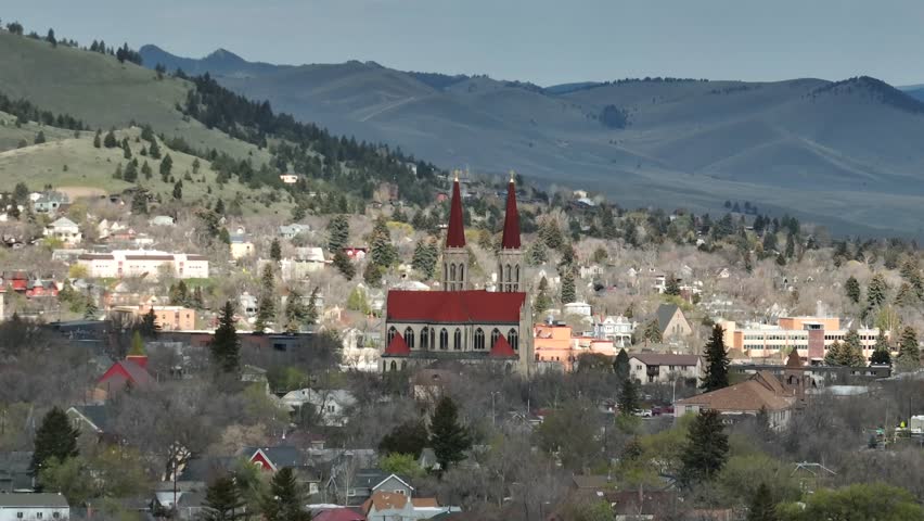 Cathedral of Saint Helena’s twin spires rise above wooded hillside homes, with historic buildings and forested mountains framing Helena’s spring skyline. Montana, USA. Aerial drone wide shot
