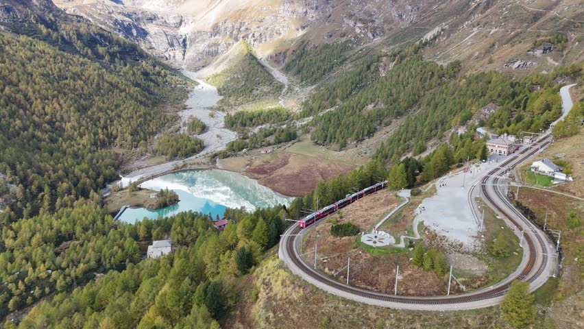 On a brisk fall day, a train  snakes through the famous curve to arrive at Alp Grüm Station in Poschiavo, Graubünden, Switzerland, while another train is parking at the station, with an aerial view overlooking Lake Palu below the receding glacier under the snow sprinkled alpine mountains in bacground