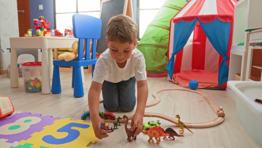 Boy kneeling and reaching with hand toward toy train on wooden floor in building playroom with tent, chair and foam puzzle mat; imaginative play joy.