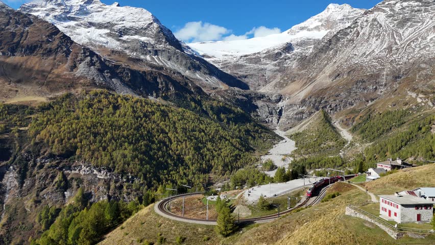 On a brisk fall day, a train of Rhaetian Railway snakes down the famous curve after departing from Alp Grüm Station in Poschiavo, Graubünden (Grisons), Switzerland, with an aerial view overlooking Lake Palu at the end of the receding glacier under the snow sprinkled alpine mountains in bacground.