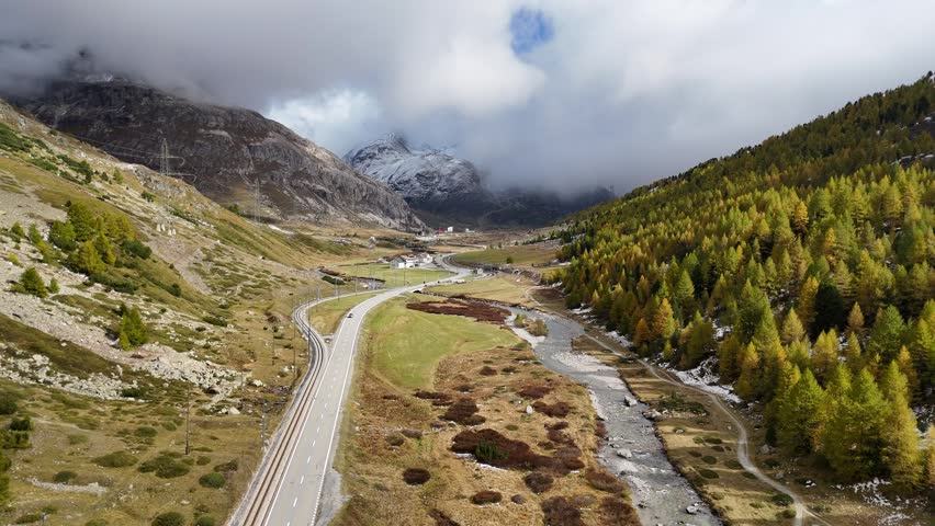 Aerial footage of a stream flowing in the valley of Bernina Pass, with the hillsides blanketed by golden larch trees on a cloudy autumn day and a highway running parallel to the railway track, in Pontresina, Grisons (Graubünden), Switzerland.