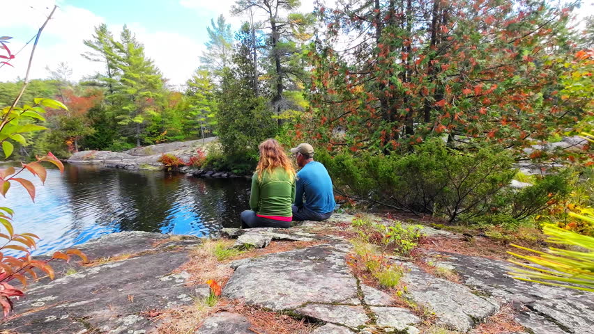 Couple sitting on a rocky ledge overlooking a lake surrounded by autumn colors in Algonquin Provincial Park, Ontario, Canada, enjoying a quiet moment in nature.