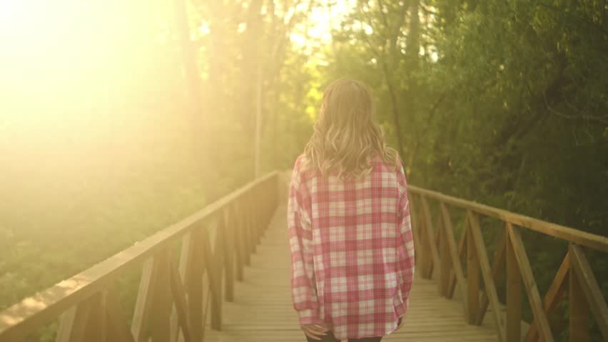 Rear view of a woman in a pink plaid shirt walking along a wooden footbridge through a green forest, with warm sunlight filtering through the trees and a calm