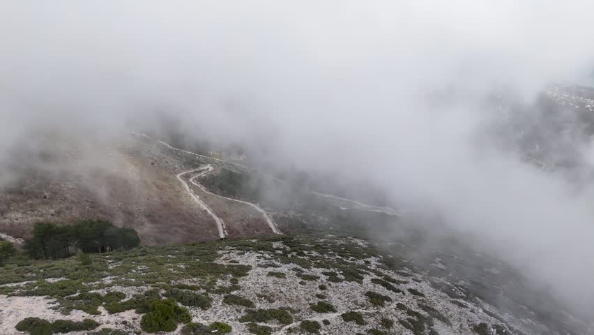 High-angle aerial shot moving forward directly through thick white fog and swirling clouds over the dramatic winding switchbacks of the Llogara Pass mountain road in Albania
