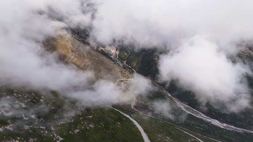 High-angle aerial shot rising upward through swirling white clouds, revealing steep rocky slopes and a winding serpentine mountain road below at Llogara Pass, Albania