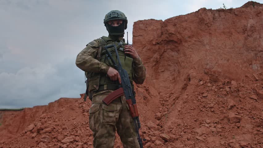 Soldier in camouflage gear aiming rifle from red desert slope with alert side profile, overcast light, secure two-handed stance, ready for training or defense duties