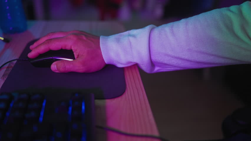 Man hand holds gaming mouse on mousepad and clicks near keyboard at studio desk under purple led lighting, visible cable and microphone stand; concentration gaming.