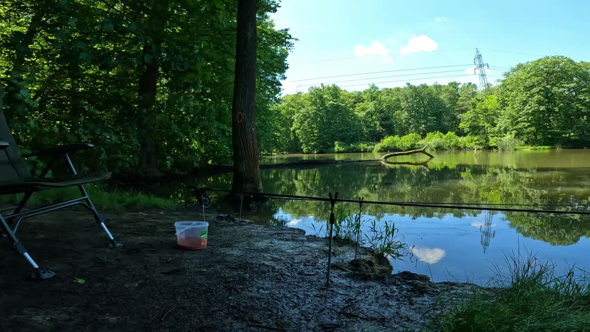 Tranquil carp fishing setup on a lake shore. Fishing rods, chair and bait bucket waiting for a bite. Peaceful outdoor recreation and relaxation.