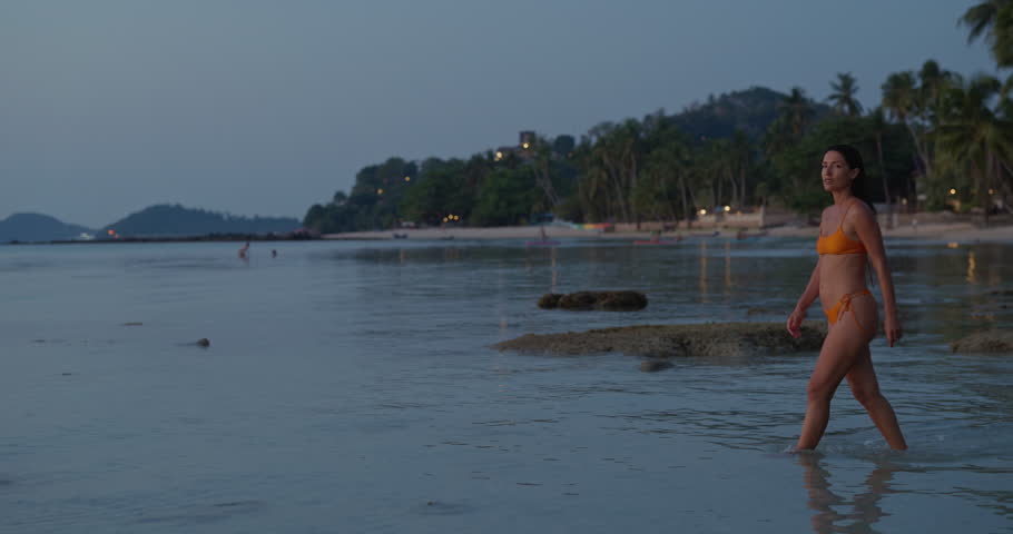 Middle Eastern woman stands in shallow sea on tropical beach during sunset dusk. Swimmer faces palm shoreline pausing calmly in clear coastal water quietly