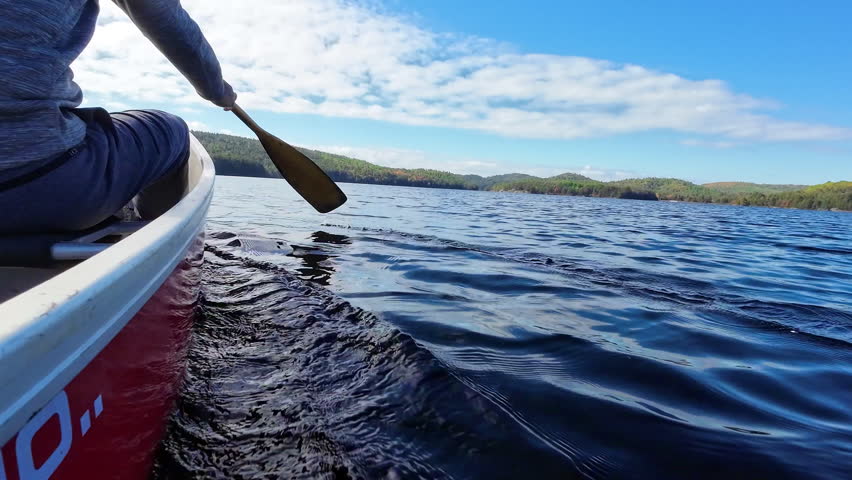 Low angle view beside a canoe with a man paddling across Grand Lake with distant hills and shoreline forest in Algonquin Provincial Park, Ontario, Canada.