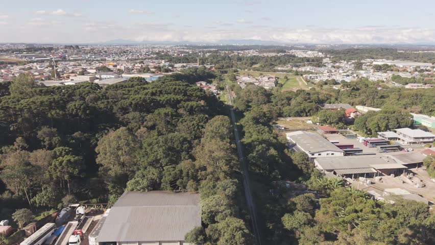Drone aerial footage following a railway corridor crossing dense urban forest and preserved green areas in the Umbara district of Curitiba Brazil with residential neighborhoods visible along the transportation route