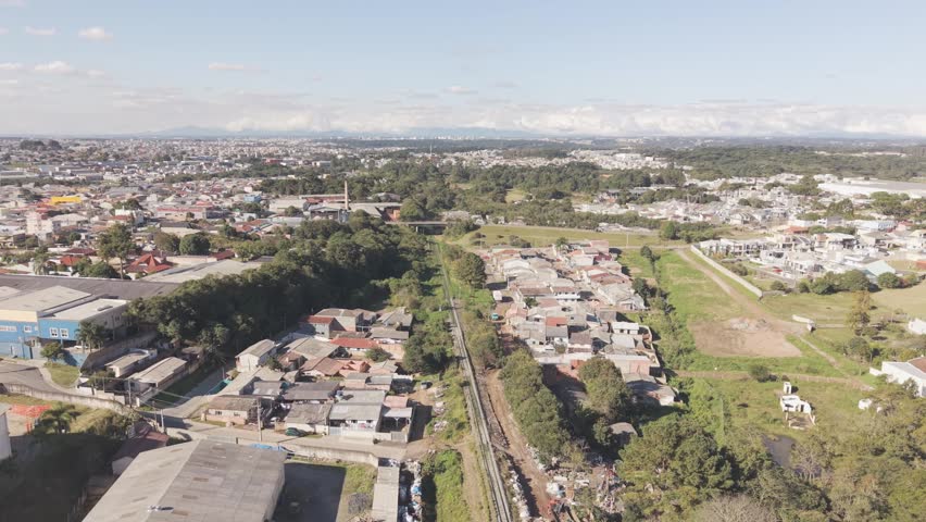 Drone aerial footage following a railway corridor approaching residential districts and roadway overpass in Curitiba Brazil surrounded by vegetation housing developments and expanding urban infrastructure