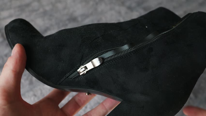 A person carefully holds and inspects a black suede heeled ankle boot in their hands. The shiny metal side zipper contrasts sharply against the dark textured fabric of the shoe.