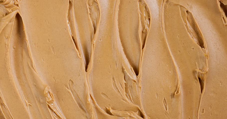 Creamy peanut butter texture background. Top view of a spoon spreading smooth peanut butter across the full frame, creating flowing lines and organic patterns. Macro food texture background.