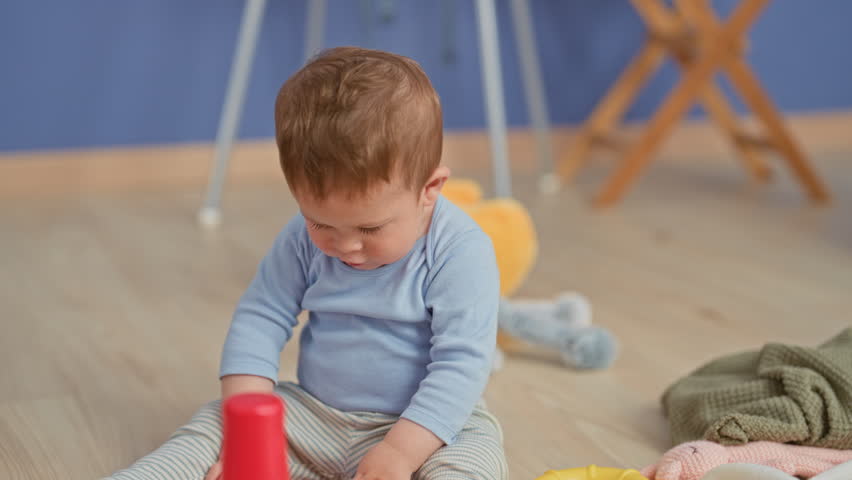 Toddler boy sitting in studio holding an orange ring toy and reaching for a red stacking cup; curiosity discovery.