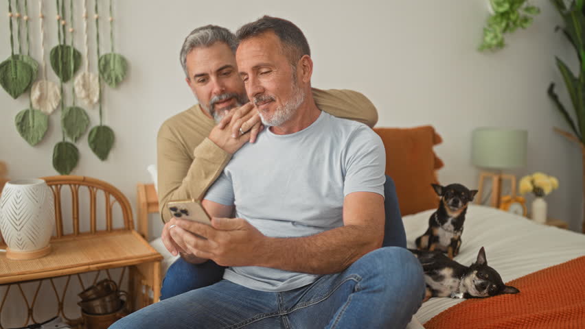 Two men laughing and kissing while one holds smartphone and rests arm around partner on bed in bedroom; intimacy.