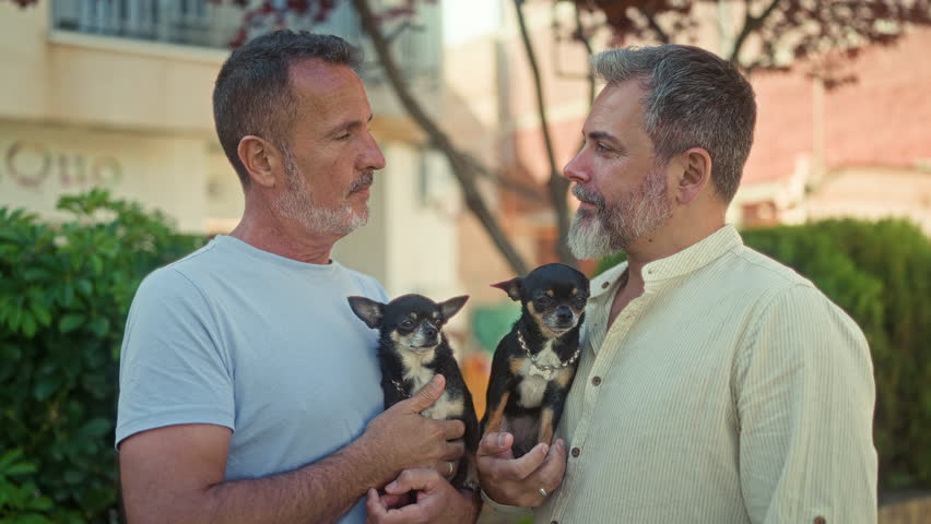 Two men hold chihuahuas, touch lips in a brief kiss and smile while facing each other in front of a residential building; affection companionship.