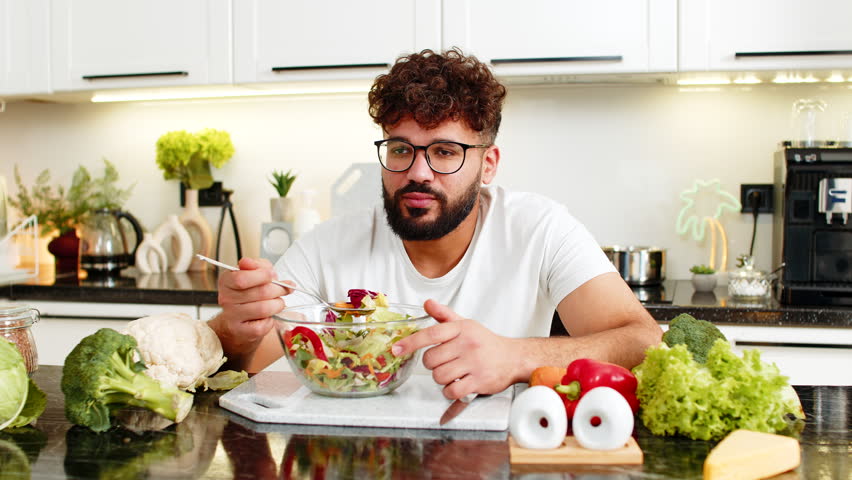 Arabian man forces salad bites in kitchen feeling tired of strict keto diet and healthy nutrition at home. Young guy bored with a diet routine craving comfort food while trying weight loss lifestyle.