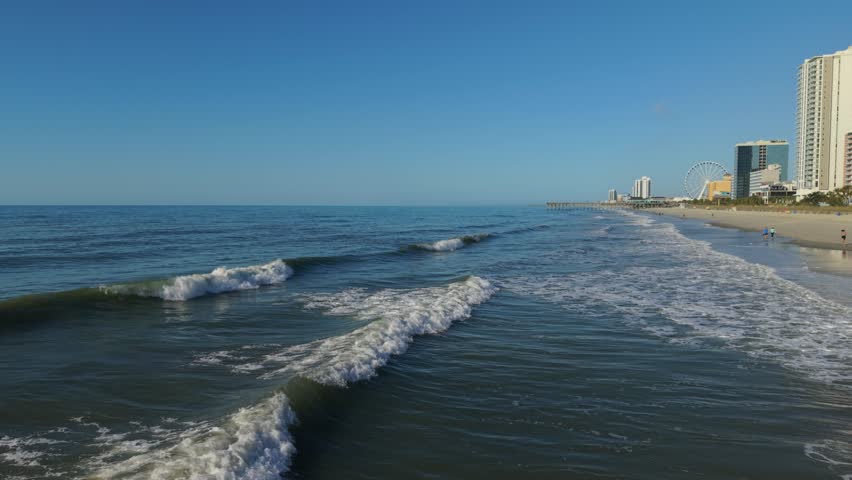 Ocean waves gently roll onto the sandy beach at Myrtle Beach, South Carolina, with coastal buildings and a ferris wheel in the background