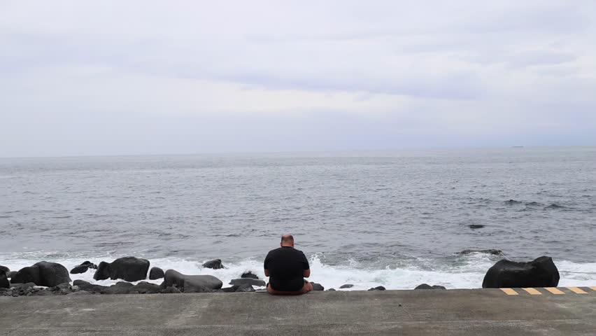 Male person sitting on concrete ledge by the ocean, enjoying serene side view of the beach with rocks and waves. Moment of relaxation during vacation or travel. Man on a beach